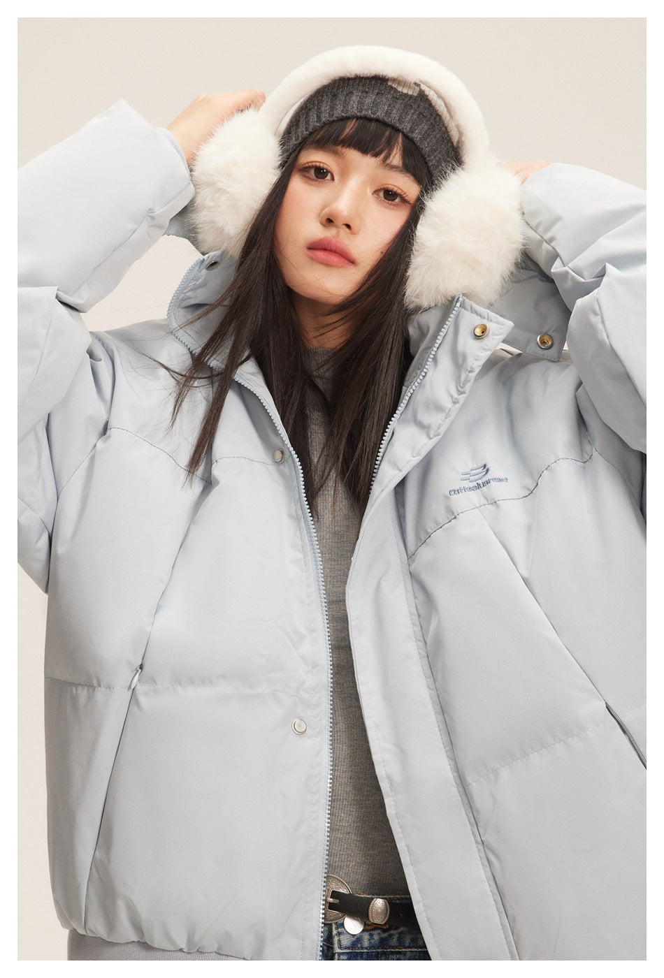 Close front view of a model wearing a light gray puffer jacket with a zip front, snaps, and chest embroidery.