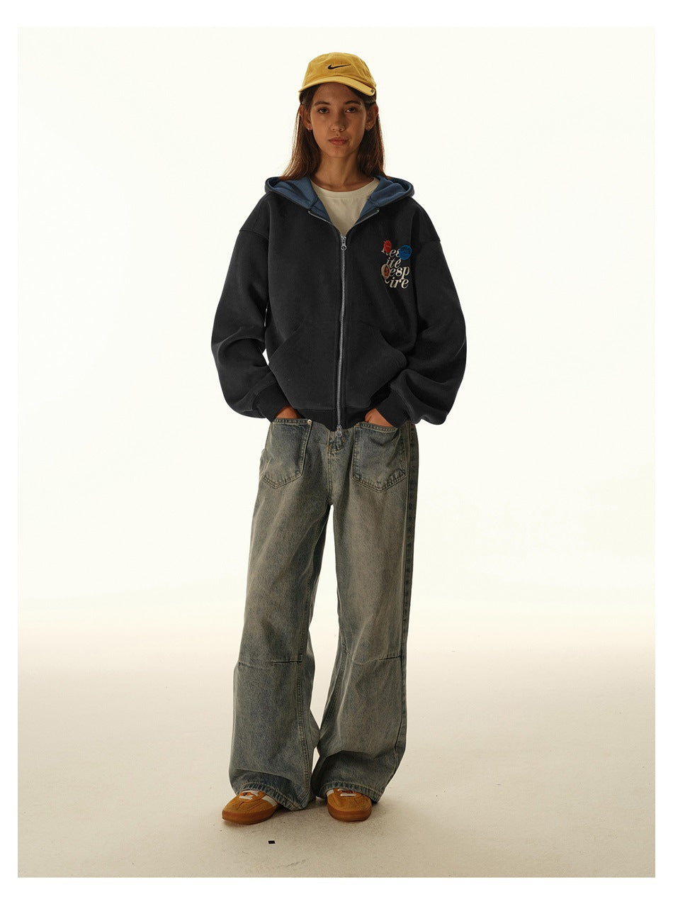 Model wearing a black zip-up hoodie with colorful chest artwork, loose gray jeans, and a yellow cap.