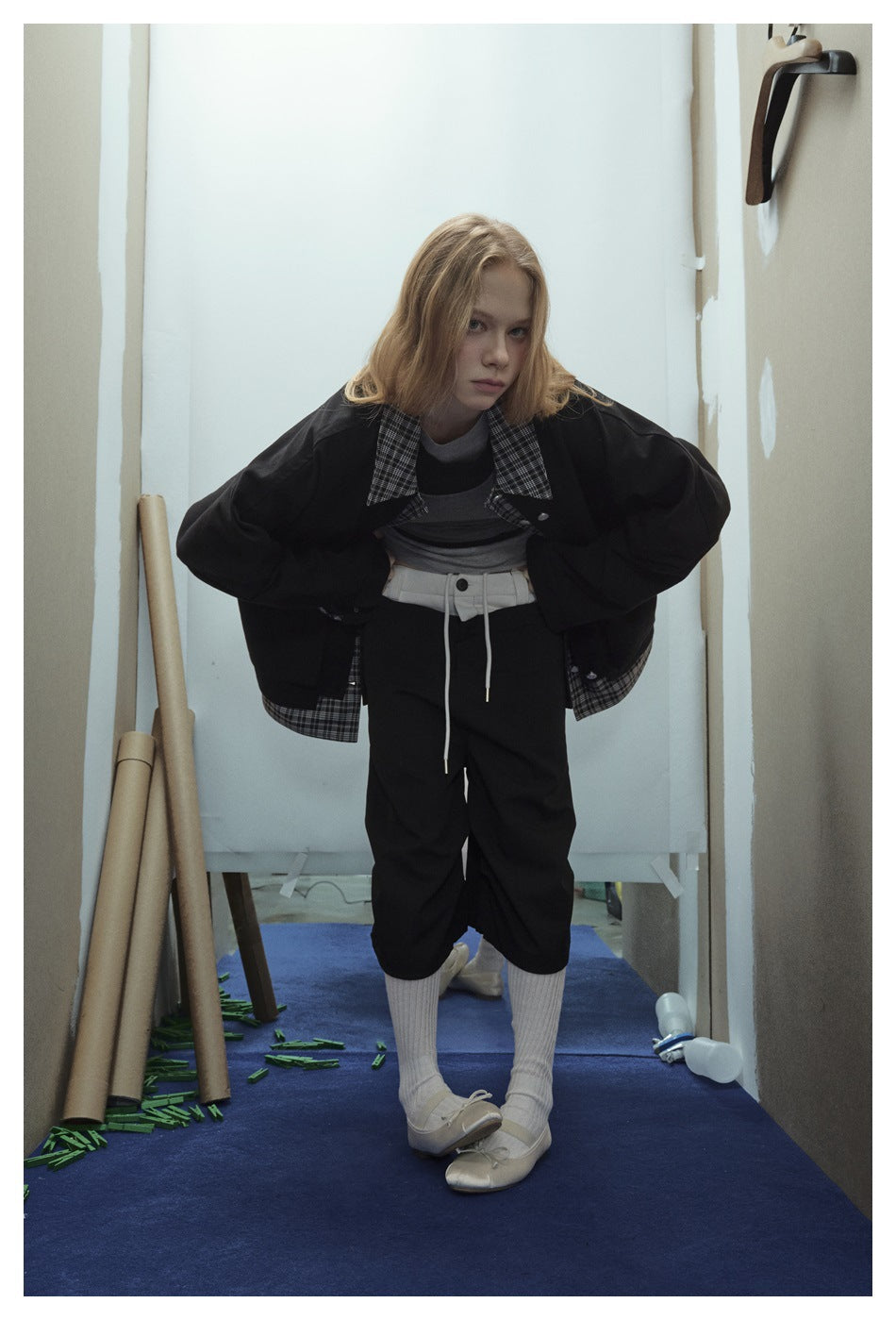 Model wearing an oversized black jacket with plaid collar and hem, layered tops, cropped black trousers, and white socks.