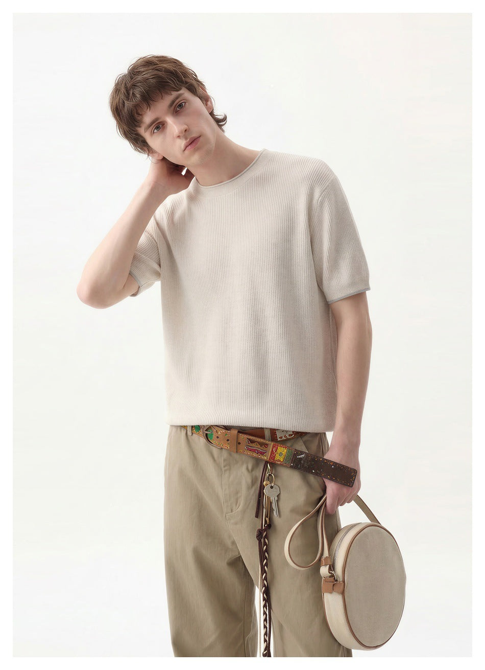 Man wearing an off-white ribbed knit short-sleeve top with khaki trousers, holding a round bag