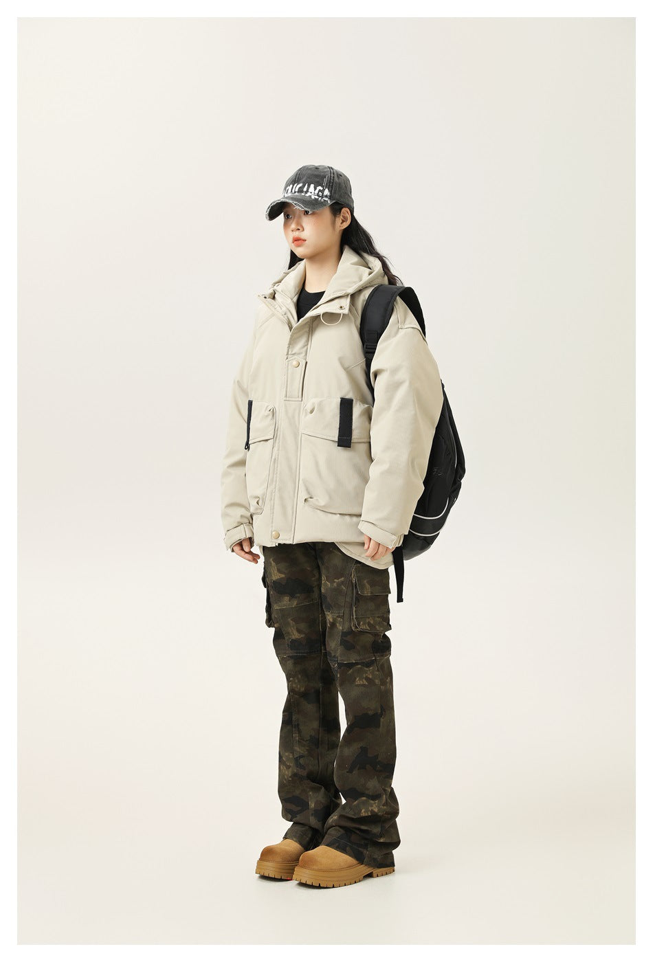 Model wearing a light beige hooded utility jacket with large flap pockets, paired with camouflage cargo pants.