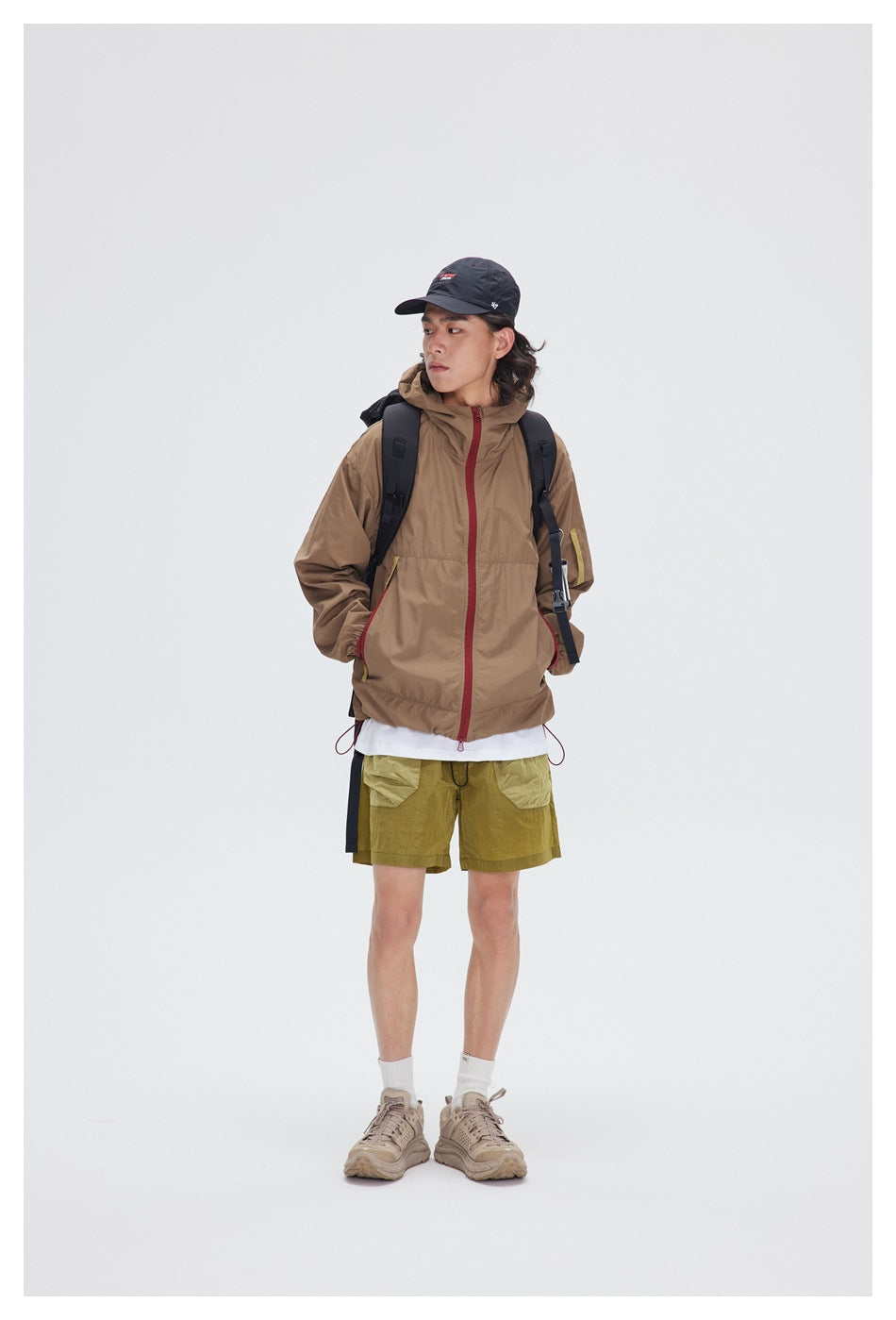 Full-length model wearing a tan hooded jacket with a red zipper and olive shorts.