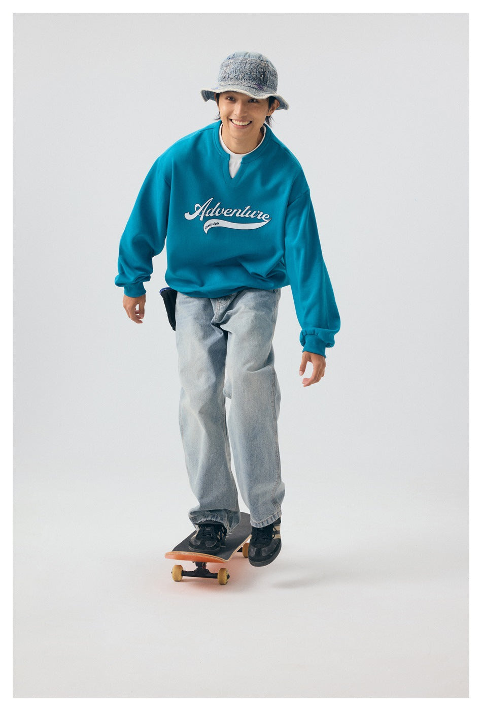 Full-length model wearing a teal Adventure graphic sweatshirt and loose jeans while standing on a skateboard.