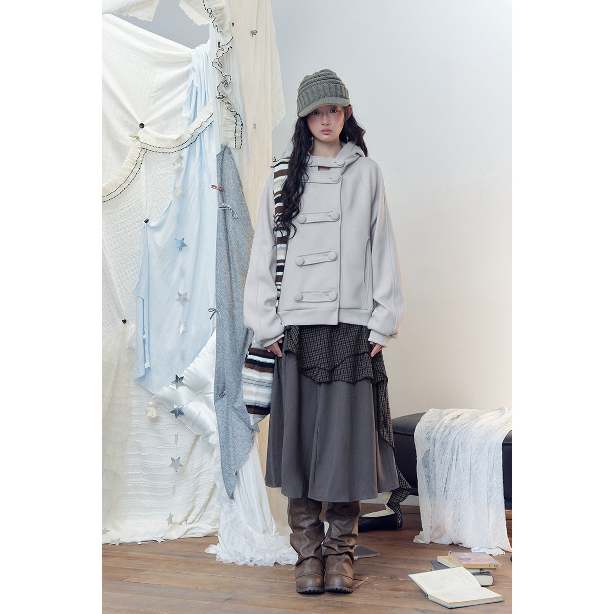 Model wearing a pale gray hooded jacket with oversized loop closures, a long gray skirt, striped scarf and knit cap.