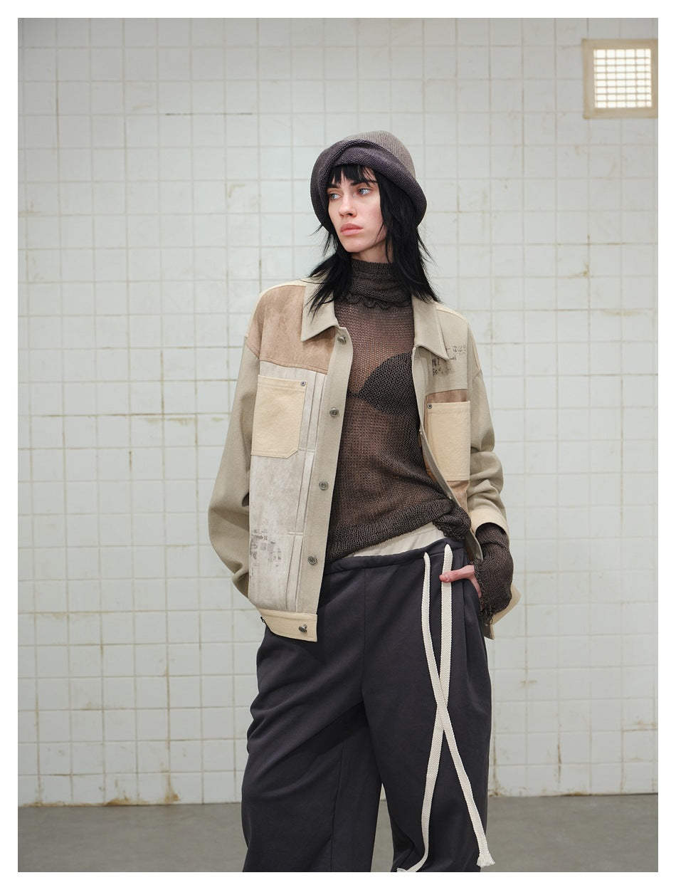 Model wearing a taupe patchwork button jacket, dark mesh top, knit hat and loose black trousers.