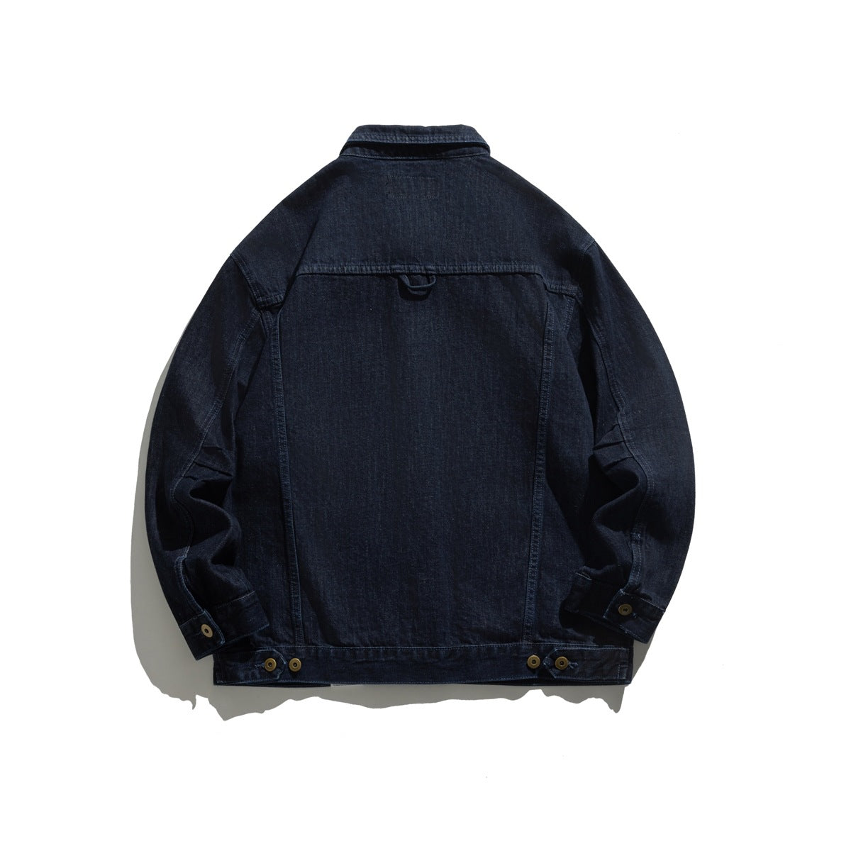 Back view of a dark indigo denim jacket laid flat, with long sleeves, adjustable buttoned cuffs, and hem tabs.