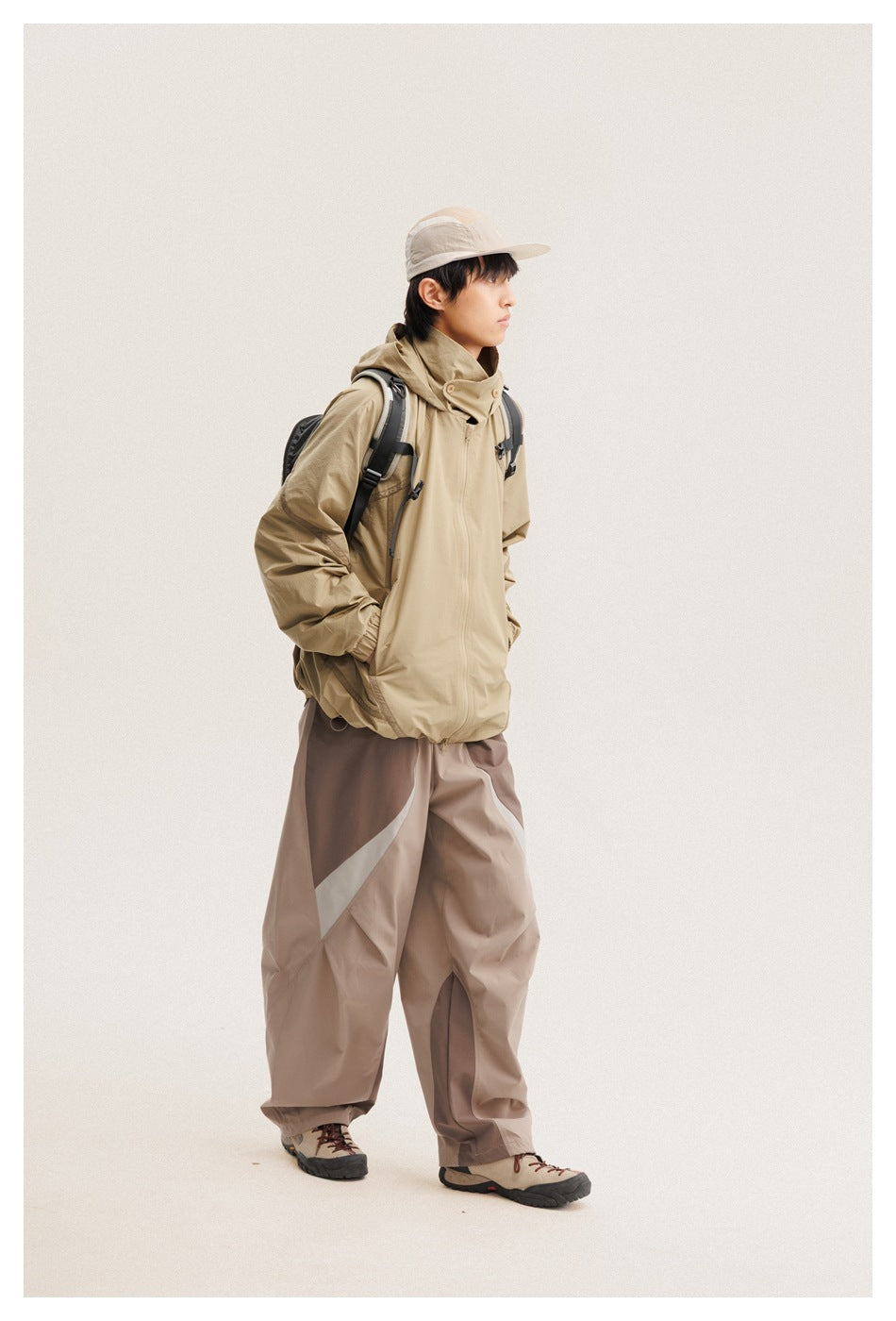 Model wearing a tan hooded jacket and loose taupe color-block pants with a dark backpack.