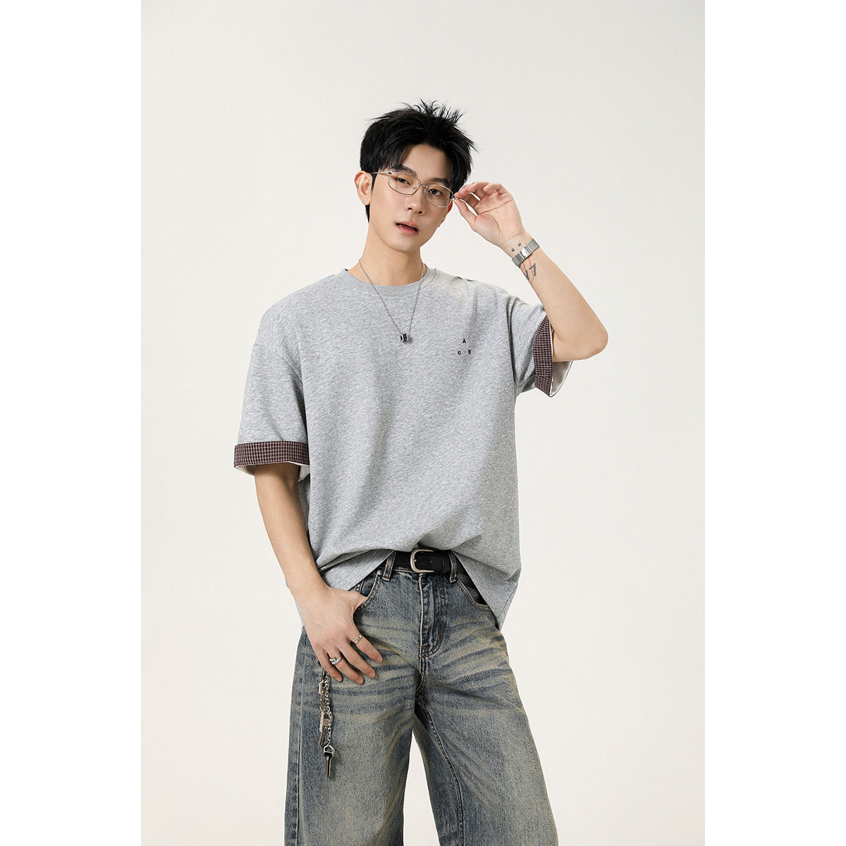 A man models an oversized light gray T-shirt with dark checked sleeve cuffs and a small chest design.