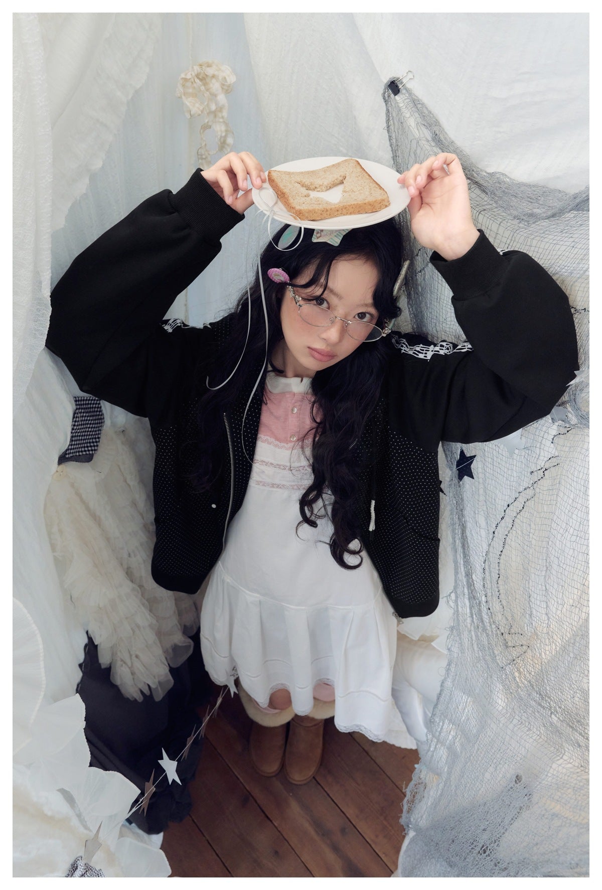 Model with raised arms wearing the black polka-dot zip hoodie over a white dress, balancing a plate with toast