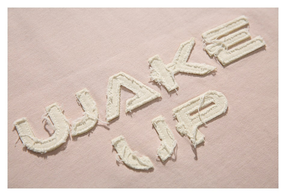 Close-up of cream textured appliqué lettering reading “WAKE UP” on pale blush-pink T-shirt fabric.