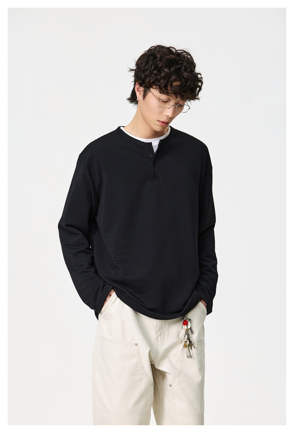Model wearing a black ribbed long-sleeve henley and cream trousers, looking down with hands in pockets.