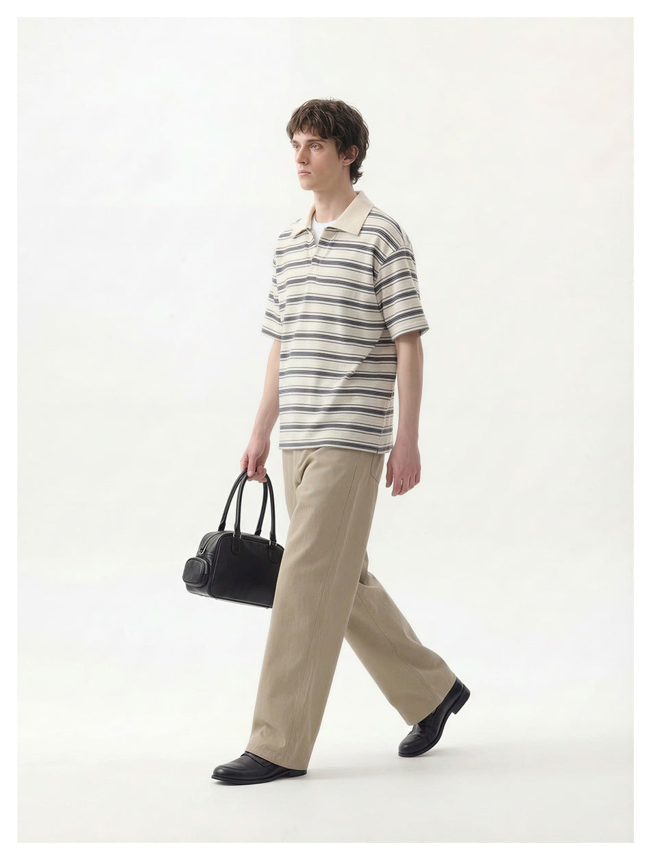 A man walks in an ivory polo with dark horizontal stripes and beige trousers, carrying a black handbag.