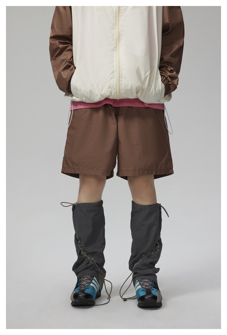Front view of a person wearing a white jacket with brown sleeves, brown shorts, and dark gaiters over blue shoes.