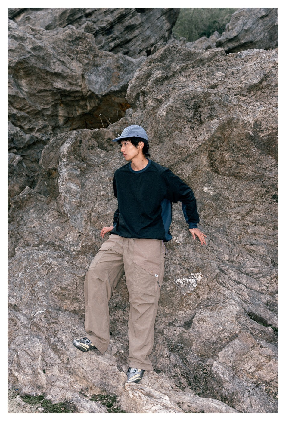Model on rocky terrain wearing a dark navy long-sleeve top, taupe cargo pants, and a gray cap.