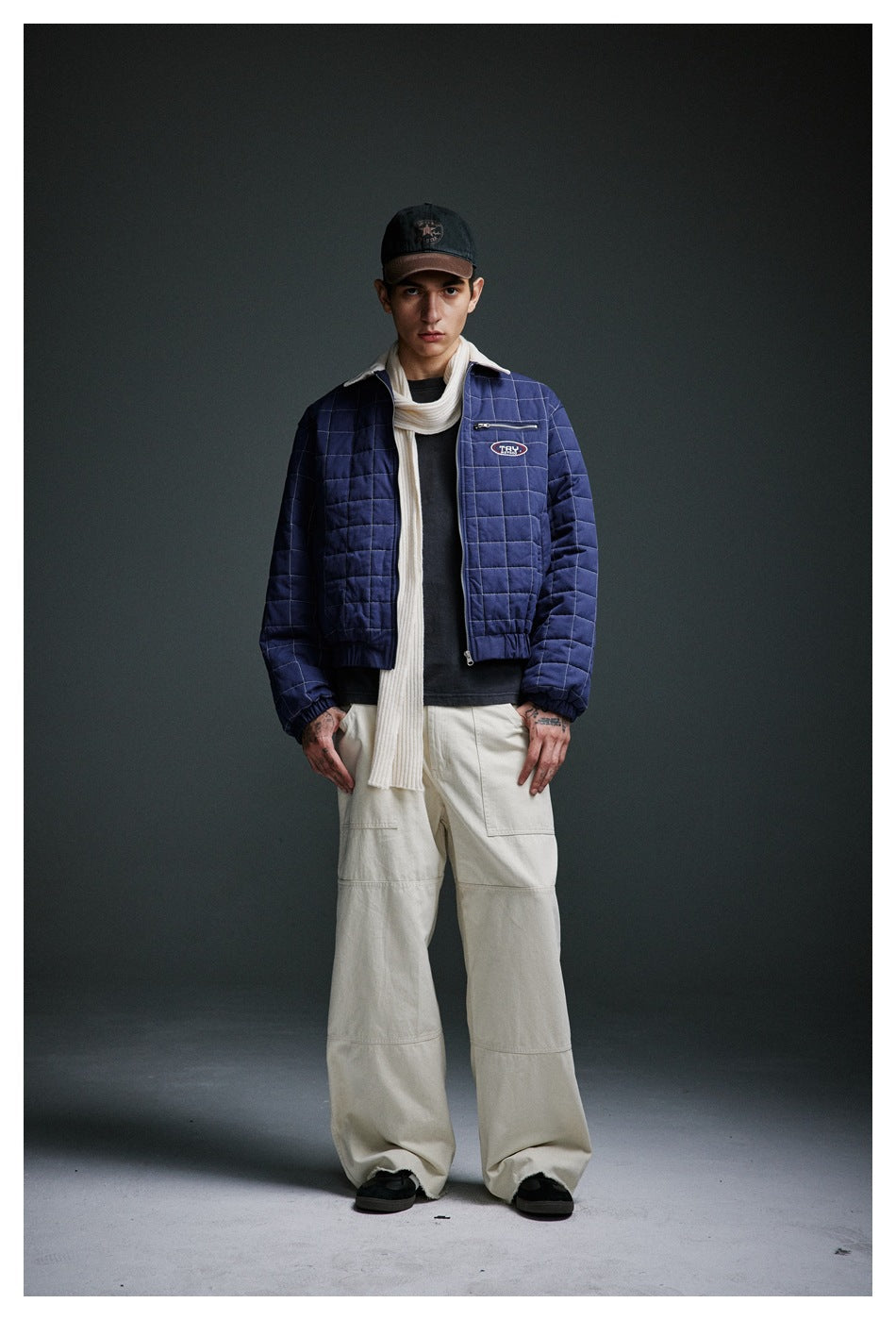 Man wearing the blue quilted jacket open over a dark tee with a cream scarf and wide cream pants