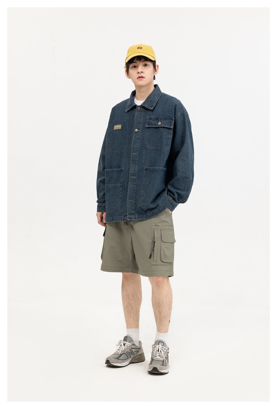 A model wears a dark blue denim chore jacket with a chest flap pocket and two large lower pockets, paired with cargo shorts.