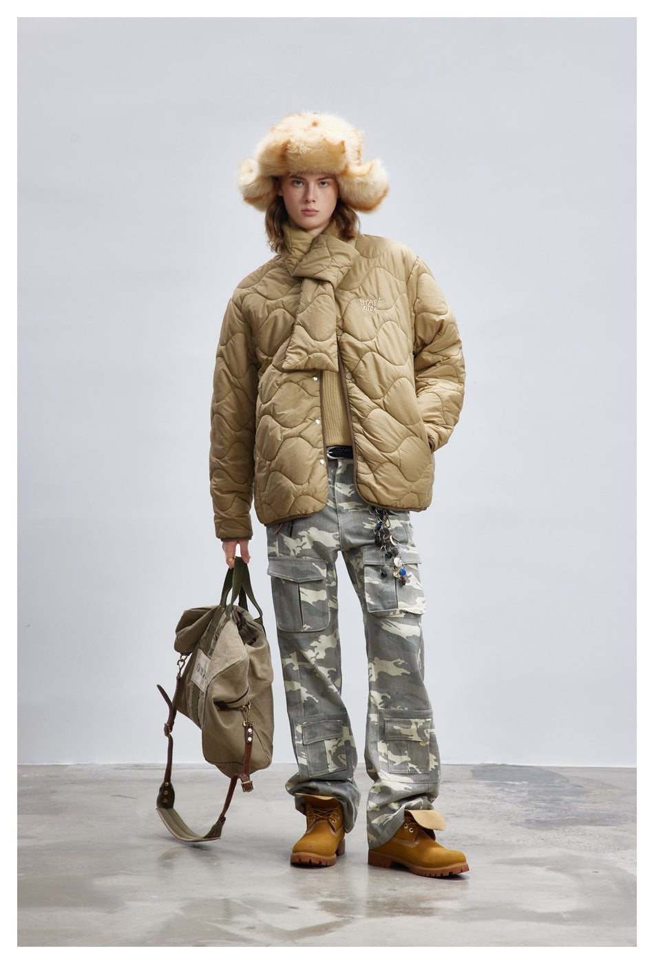 Model wearing the khaki quilted jacket with scarf piece, camouflage cargo pants, boots and a fluffy hat, holding a bag