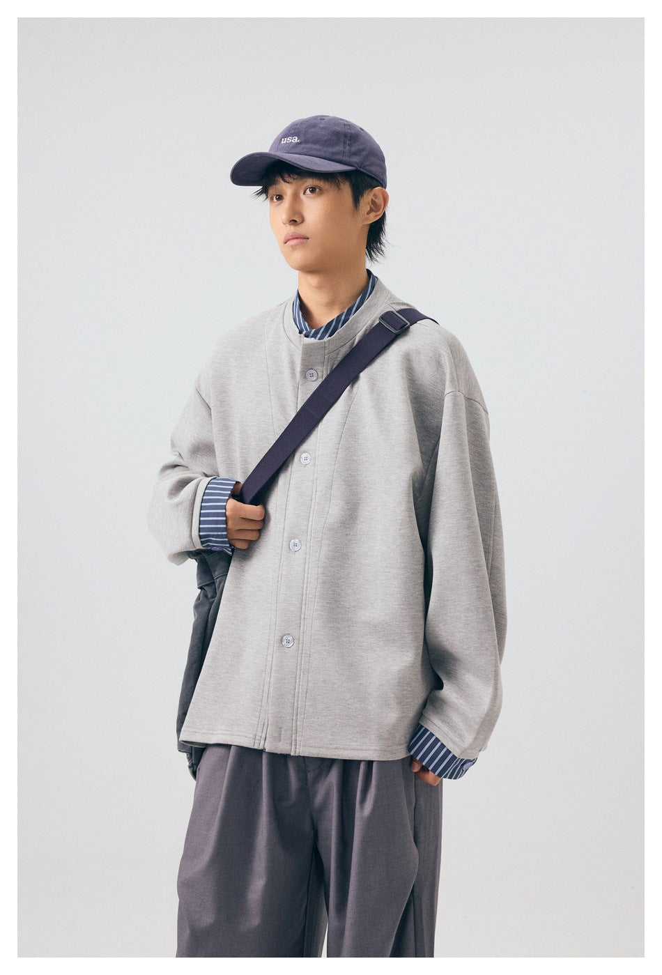 Model wearing a light gray button-front jacket with striped shirt cuffs and loose charcoal trousers, carrying a shoulder bag.