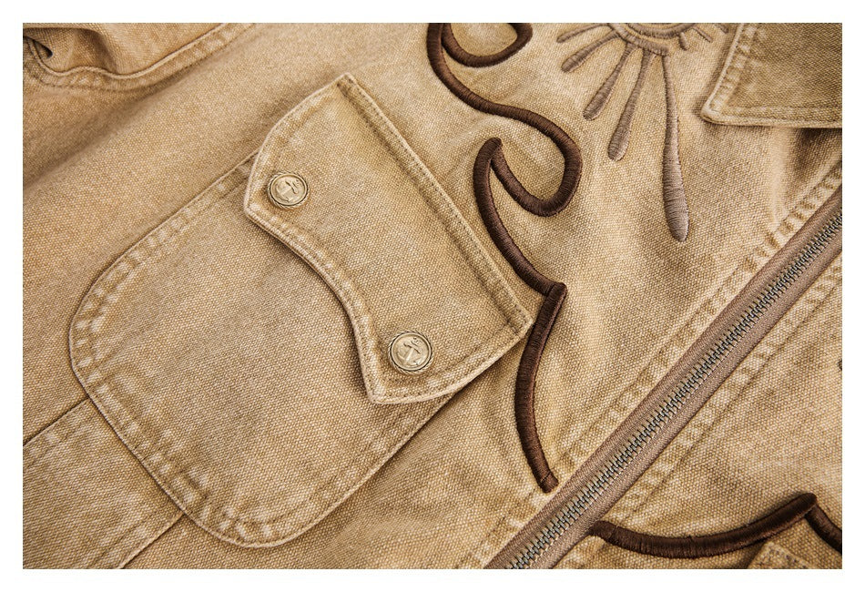 Close-up of a tan jacket's flap pocket with two buttons, dark embroidered curves, and a diagonal zipper.