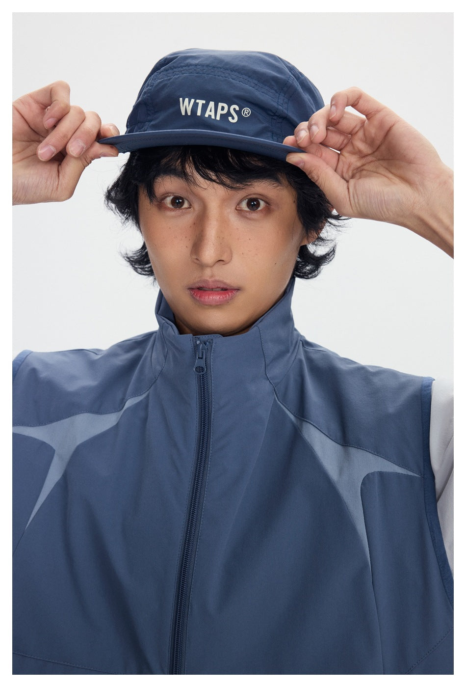 Model wearing a navy WTAPS cap and slate-blue zip-front vest with contrasting geometric panels.
