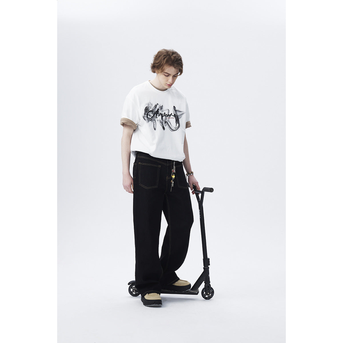 Model wearing a white graphic T-shirt with tan rolled cuffs and loose black trousers, standing beside a scooter.
