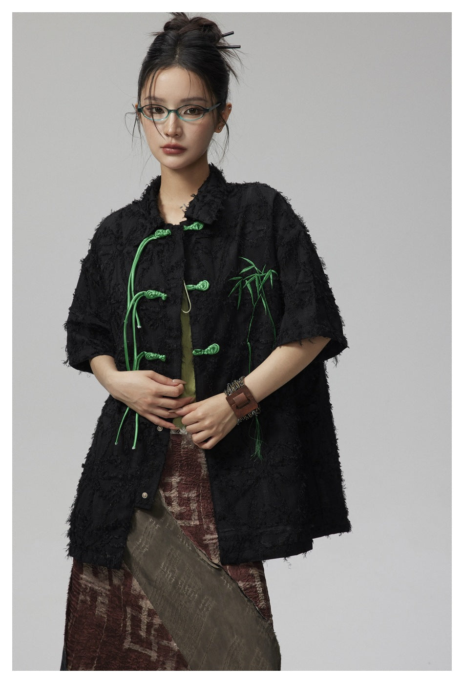 Model wearing a black textured short-sleeve jacket with green frog closures and bamboo embroidery, hands held at the front.