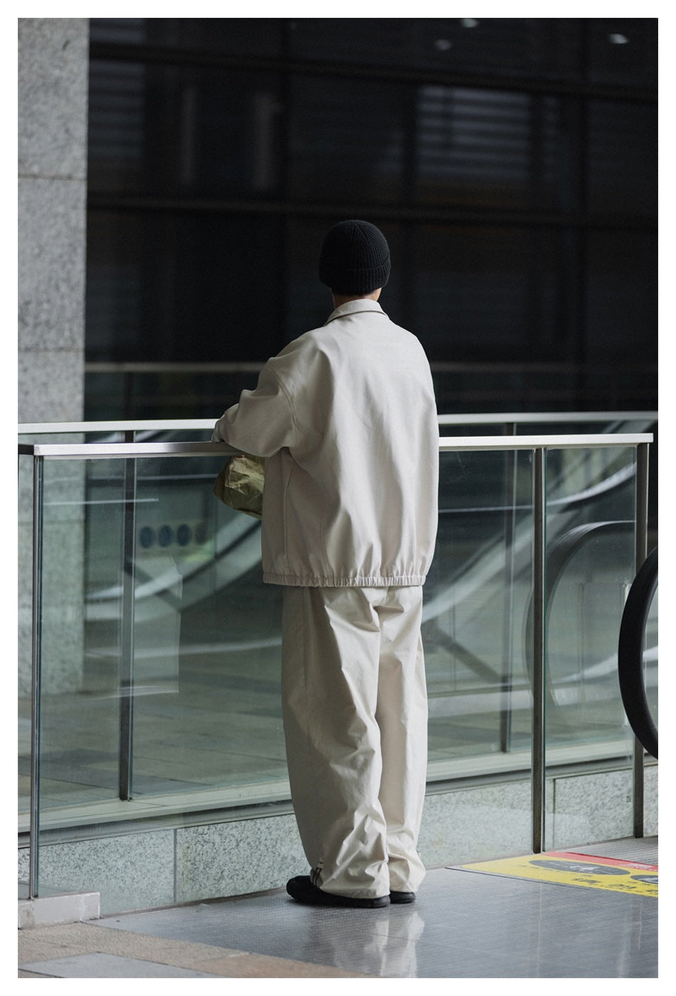 Rear view of a model in a light beige jacket with an elastic hem, matching loose trousers, and a black beanie.