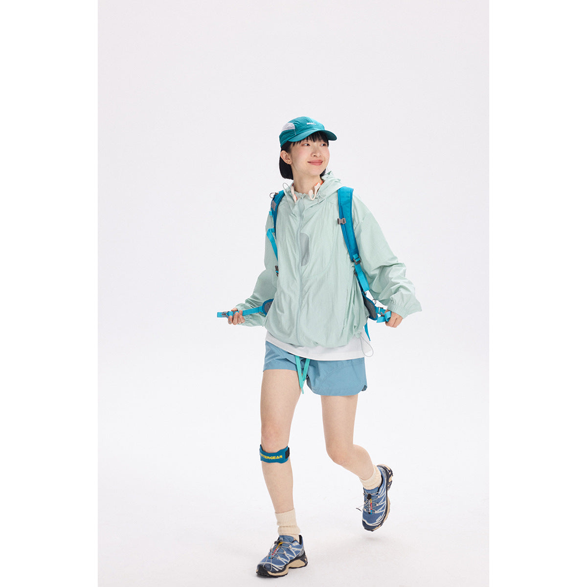 Model wearing a pale mint hooded jacket, blue shorts, teal cap, and backpack against a white background.