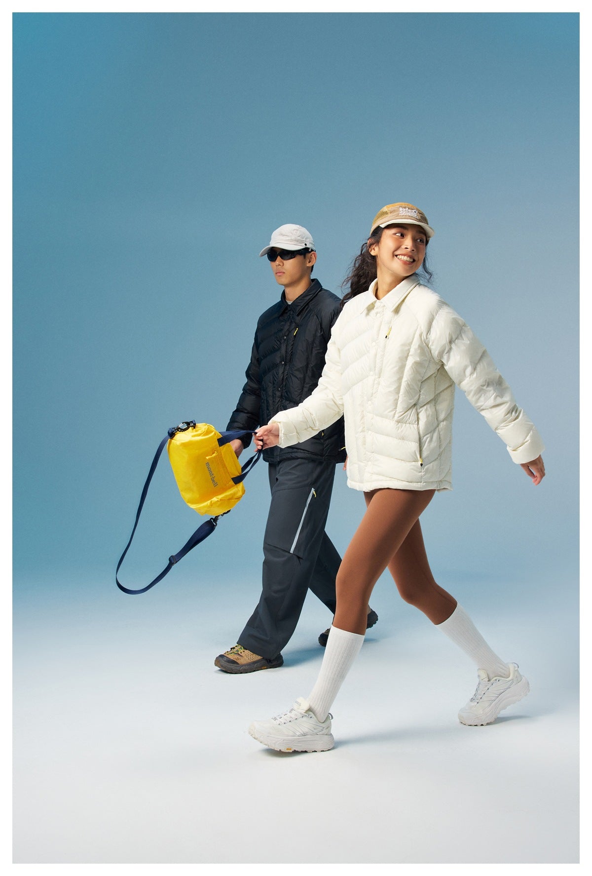 Two models wearing cream and black puffer jackets; the woman carries a bright yellow bag.