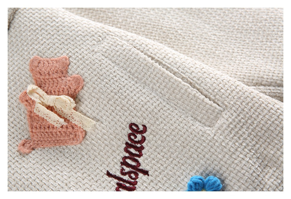 Ivory textured fabric with pink knitted appliqué, lace trim, embroidered lettering, and part of a blue flower.