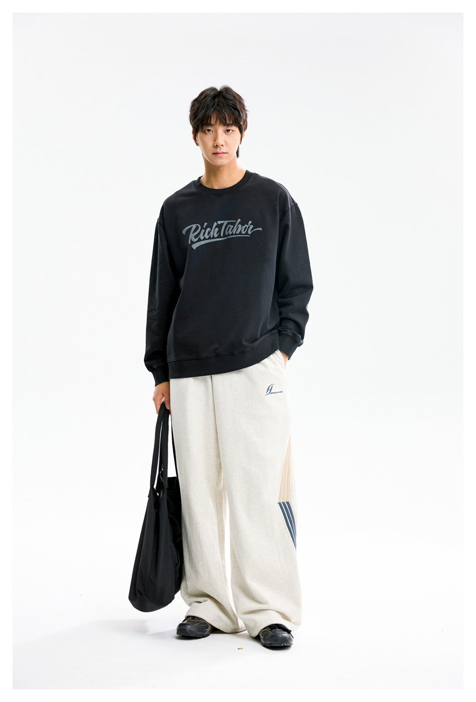 Full-length model wearing a black script-logo crewneck sweatshirt and loose off-white pants.