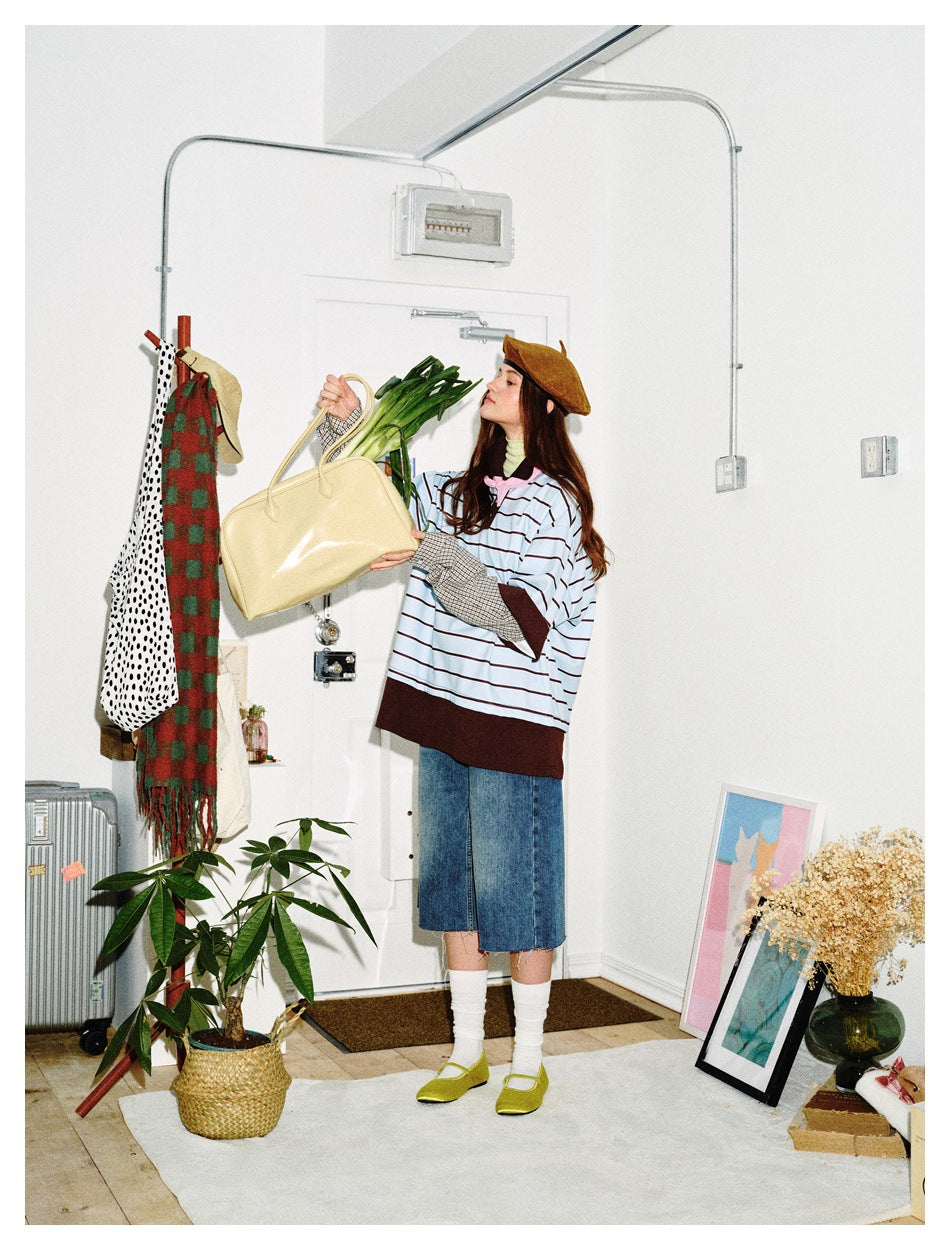 Model wearing a light blue striped top with brown trim and plaid sleeves, holding a yellow bag and leafy greens.