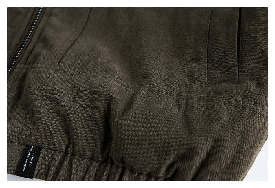Close-up of the olive-brown jacket's front pocket seam, zipper, and gathered hem with a small black label.