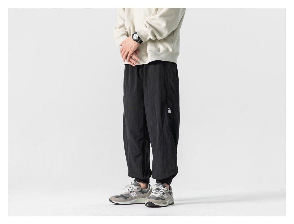 Model wearing loose black jogger pants with elastic cuffs and a small white thigh emblem, shown at a slight front angle.