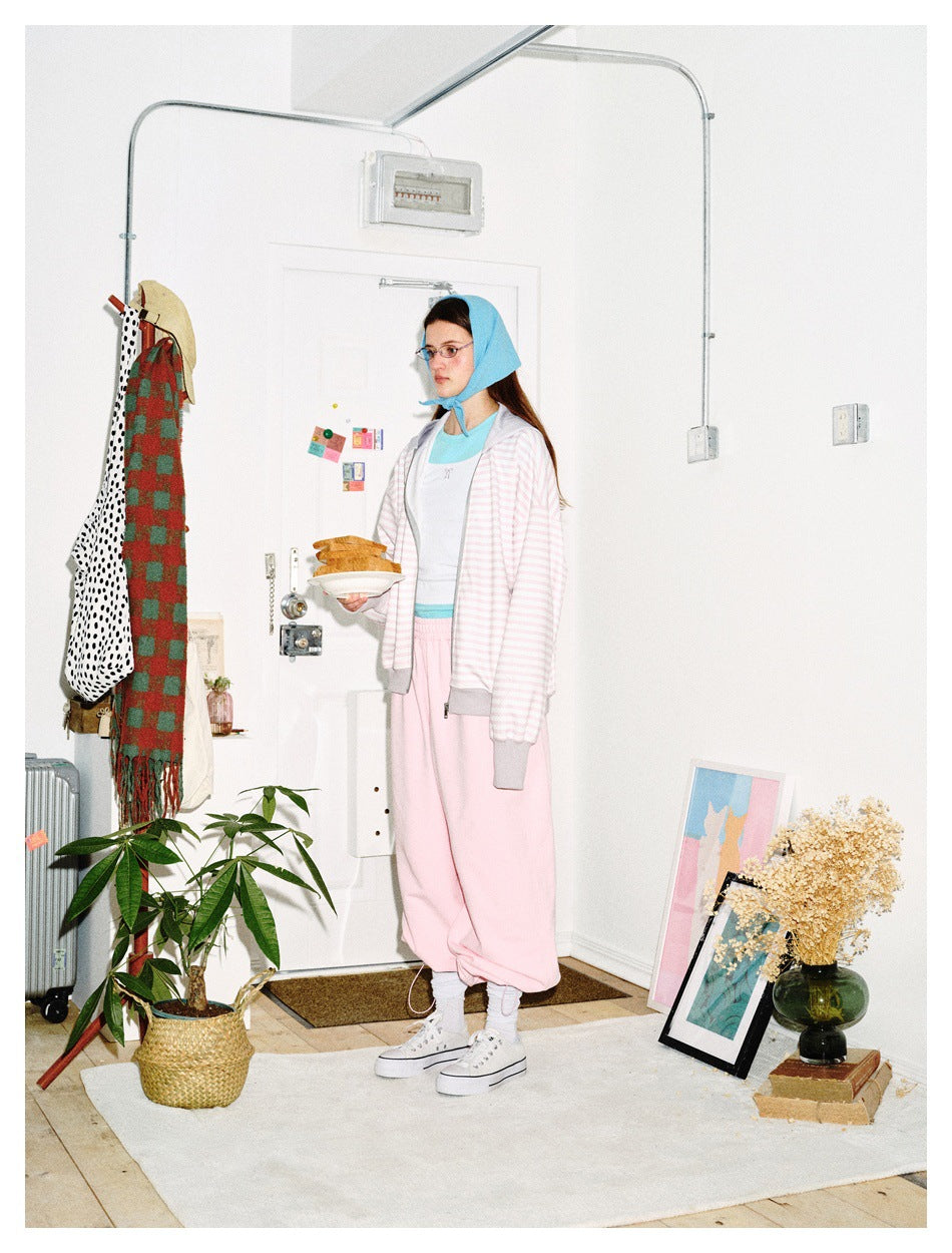 Model wearing a pale pink-and-white striped zip hoodie with gray trim and pink joggers, shown in profile in a room.