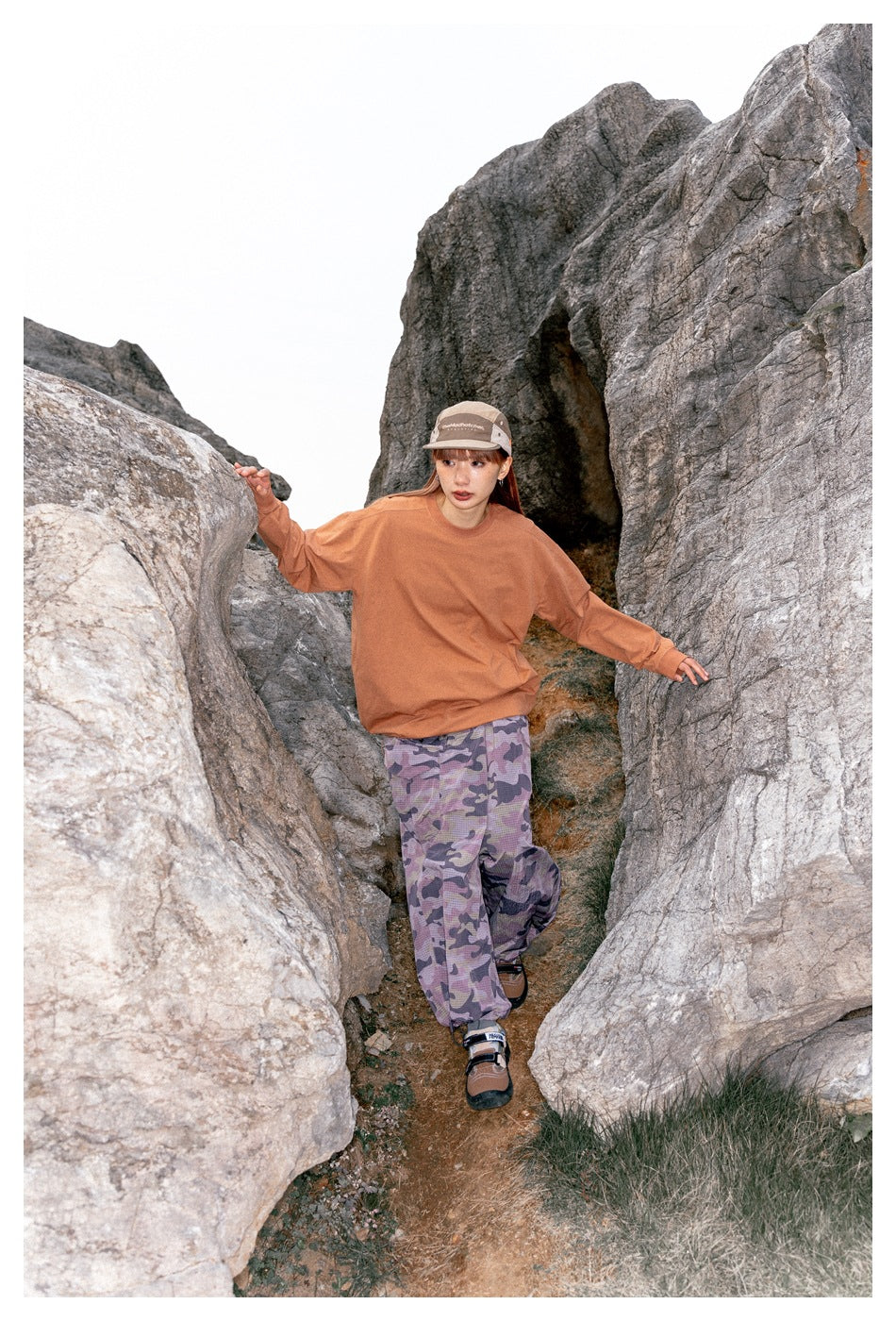 Model in a rust-orange sweatshirt and purple camouflage pants stepping through a rocky passage.