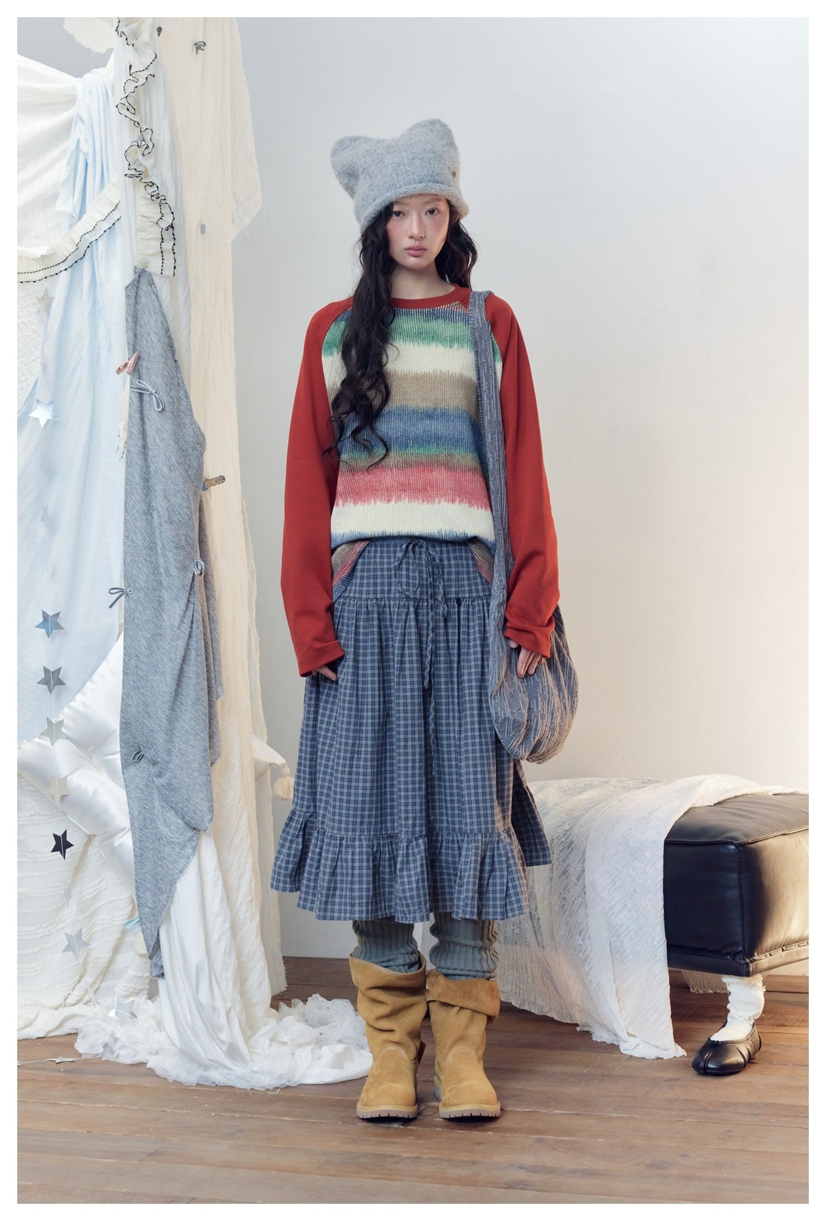 Model wearing a cream multicolor striped sweater with red sleeves, a blue plaid tiered skirt, gray hat and tan boots.