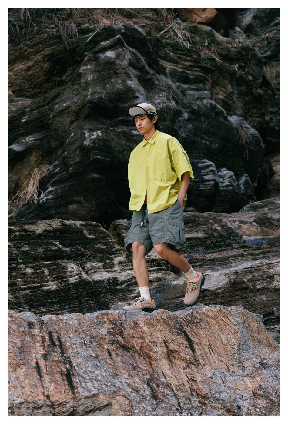 A man walks over rocks in a lime-yellow short-sleeve shirt and gray cargo shorts.