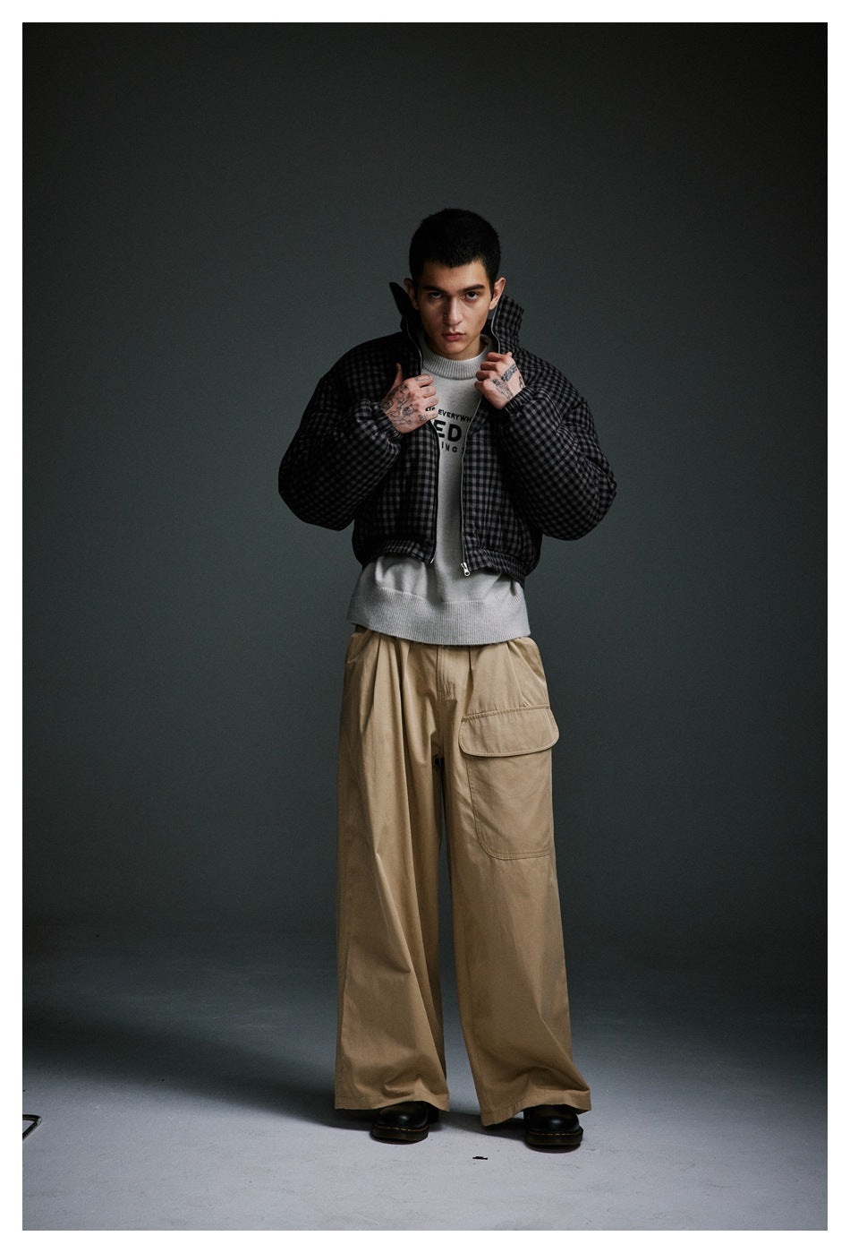Model wearing an open charcoal checked padded jacket over a white sweater and wide tan cargo trousers.