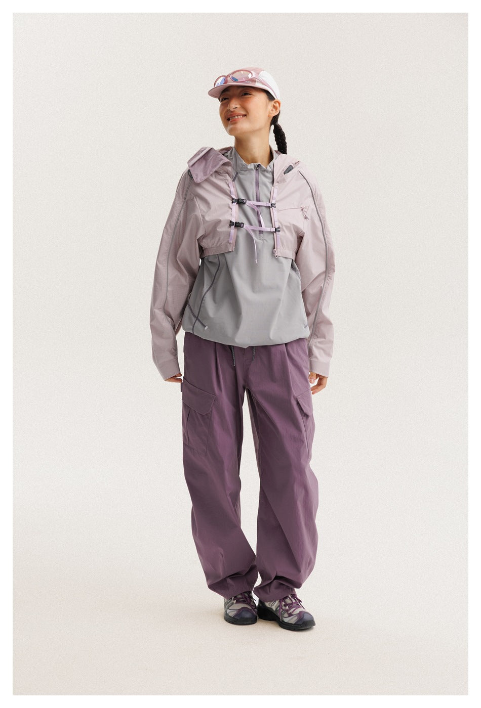 Person wears a cropped pale mauve jacket over a gray pullover, with plum cargo pants and a pink cap.