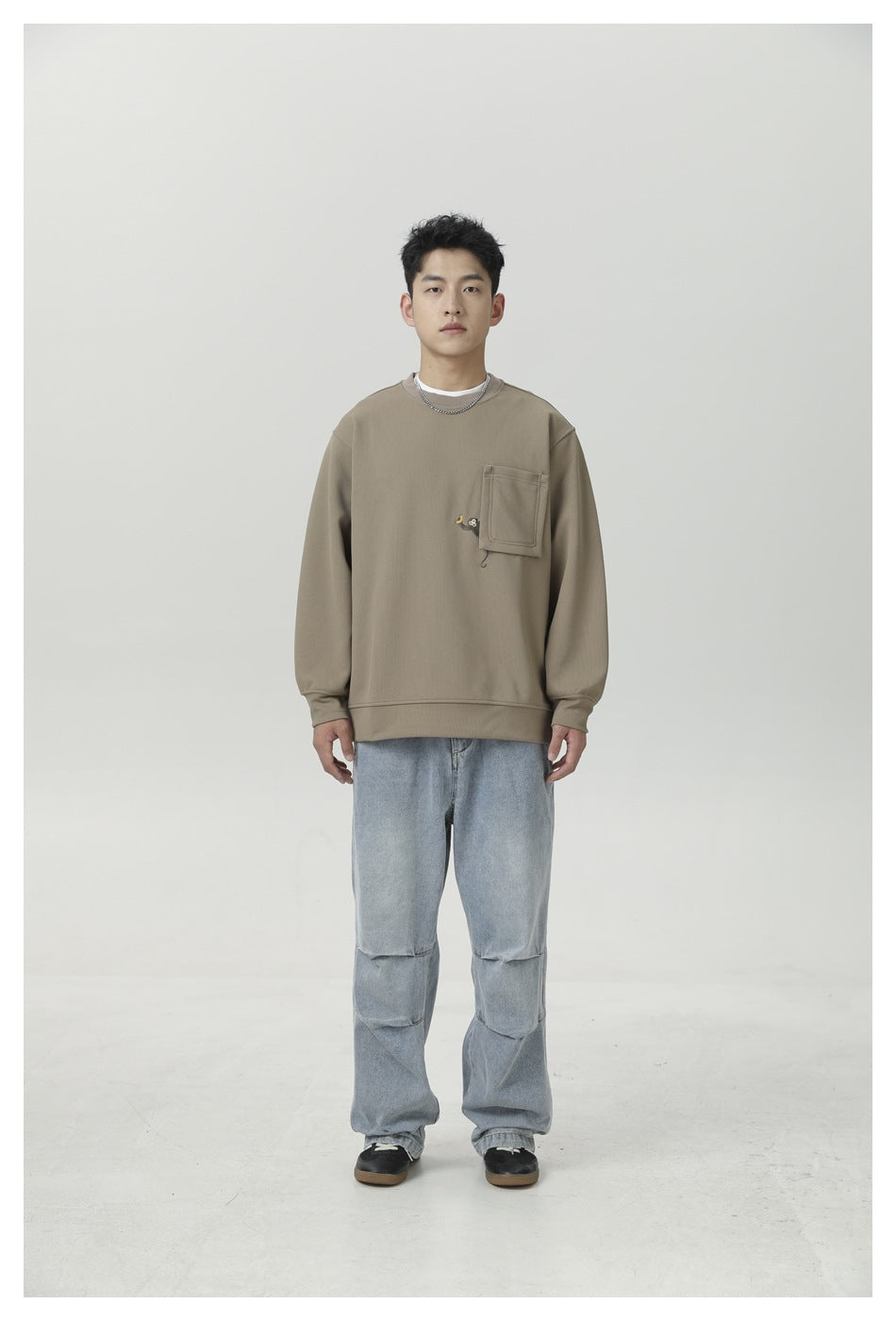 Model wearing light brown crewneck sweatshirt with chest pocket, light wash baggy jeans and dark sneakers, front view