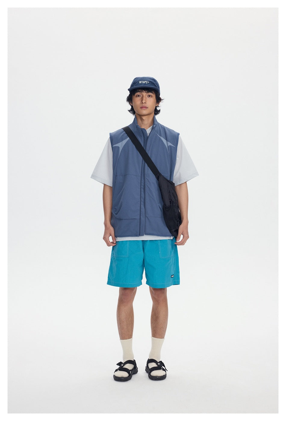 Model wearing a blue zip-up stand-collar vest over a light tee with aqua shorts and a black crossbody bag, front view