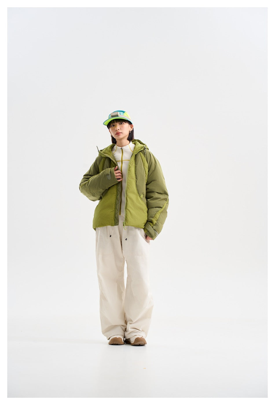 Full-length model wearing an open olive and lime-green hooded jacket over a white top and loose white trousers.