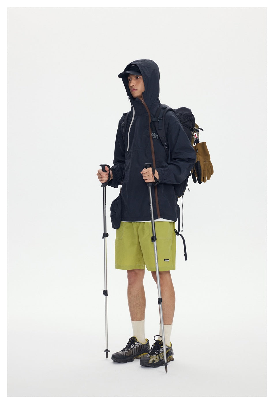 Model wearing a dark hooded jacket and lime-green shorts with a hiking backpack and trekking poles.