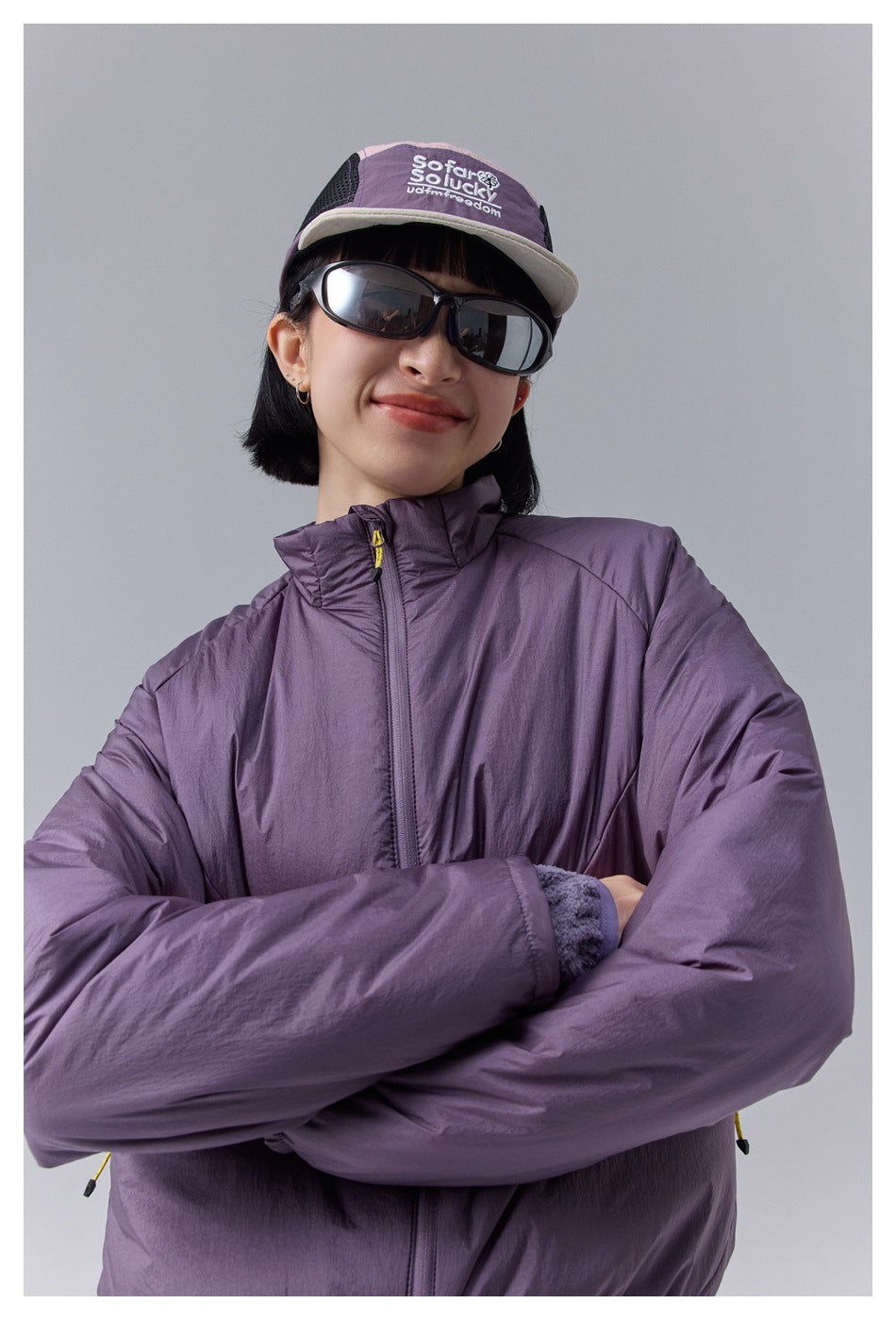 Model wearing a plum padded zip jacket with a high collar, crossed arms, sunglasses, and a cap with an English slogan.