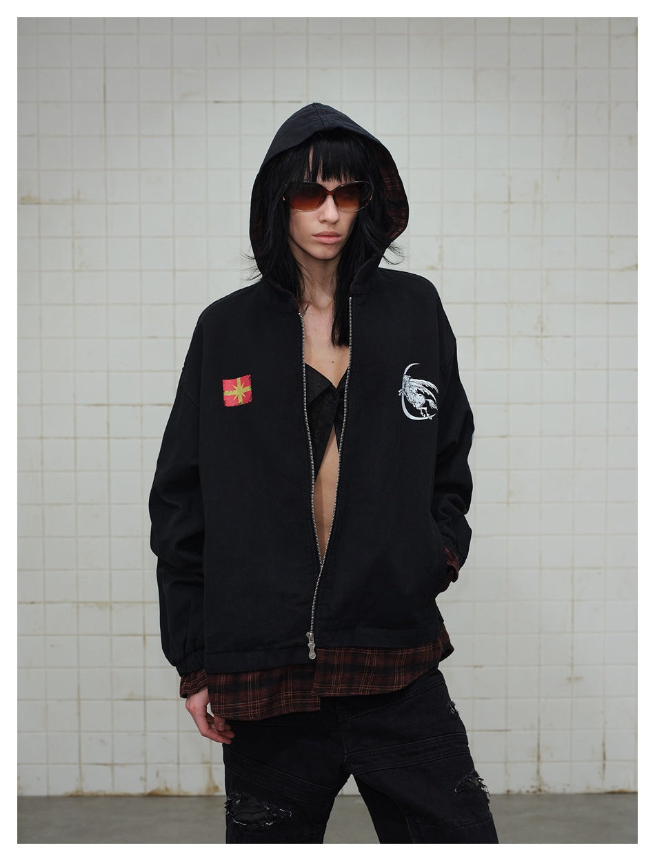 Front view of a model in a black hooded zip jacket with chest graphics and a red plaid layer visible at the cuffs and hem.