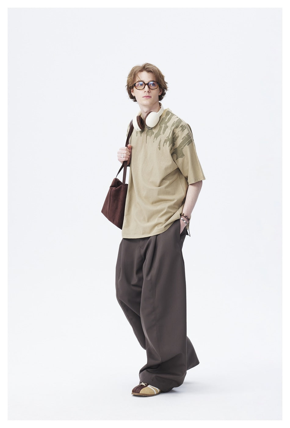 Model wearing khaki green printed T-shirt with wide brown trousers, headphones and a brown tote bag, full-length view