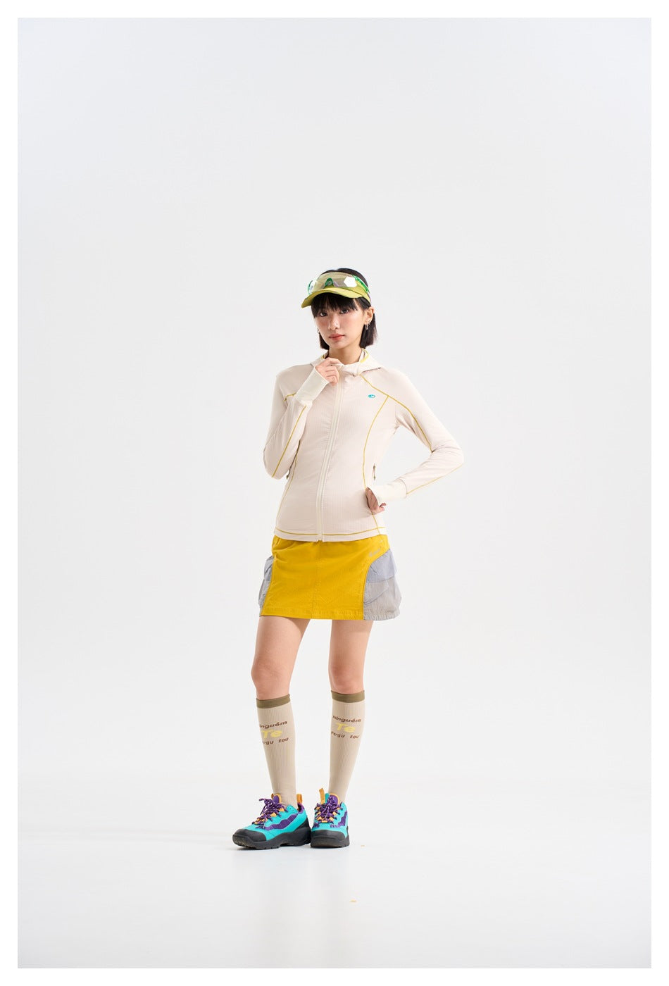 Model wearing a cream zip-up hooded jacket with yellow piping and a yellow-and-gray skirt, cap, socks and colorful hiking