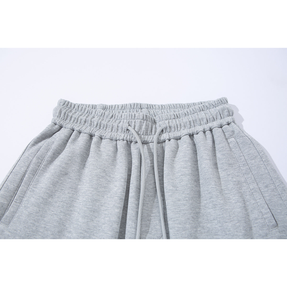 Close-up of light gray sweatpants with a gathered elastic waistband, drawstring, and slanted side pockets.