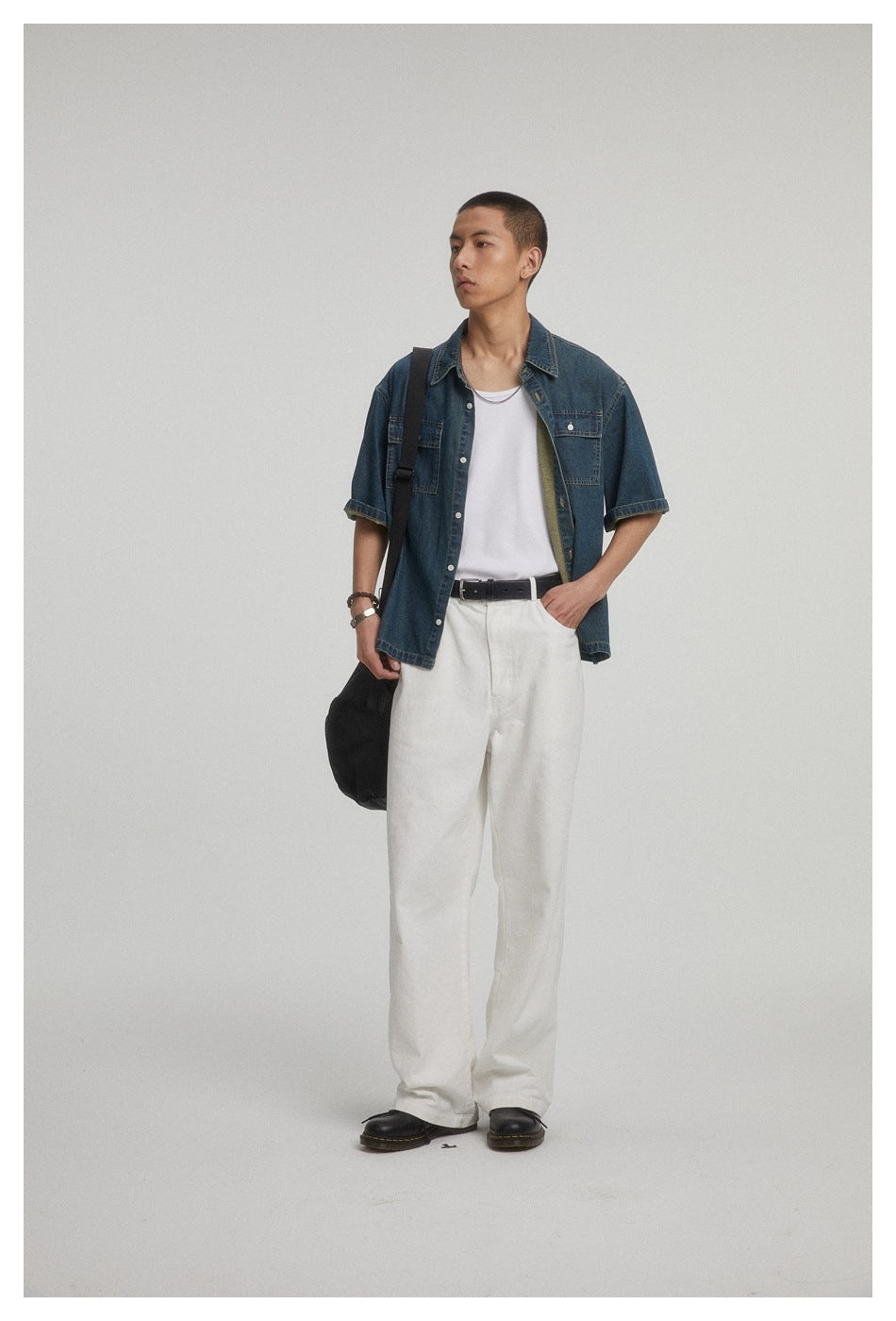 Model in a dark blue short-sleeve denim shirt with two chest pockets, white trousers and a black shoulder bag.